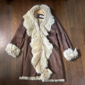 May include: A brown coat featuring a long, cream-colored faux fur collar that extends down the front, matching faux fur trim on the sleeve cuffs, and a faux fur hemline. The coat has a smooth, matte finish and is displayed on a wooden hanger.