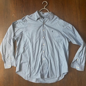 May include: Light blue button-down shirt with a small checkered pattern. The long-sleeved shirt has a button-down collar and cuffs. A small embroidered logo is visible on the chest. The shirt is hanging on a white hanger.