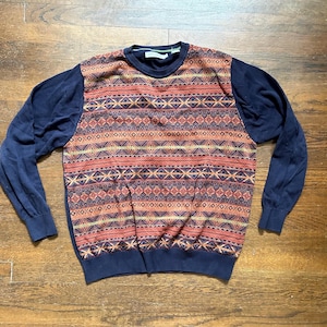 May include: A navy blue sweater with a geometric pattern in shades of red, orange, and yellow across the chest. The sleeves are solid navy blue, and the sweater has a crew neck. The garment is lying flat on a wooden surface.