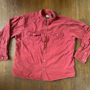 May include: A long-sleeve button-down shirt in a muted red color. The shirt features a pointed collar, two chest pockets with buttoned flaps, and a front button placket. The cuffs are also buttoned, and a Bass Pro Shops logo tag is at the neck.