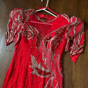 May include: A red formal dress with silver and pearl embellishments. The dress features a V-neck, short sleeves, and intricate sequin and bead detailing. The dress is hanging on a white hanger.