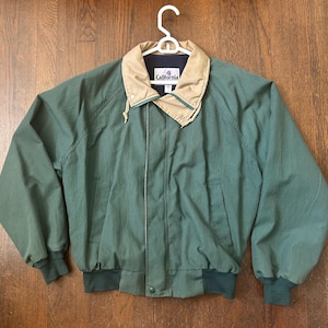May include: A green jacket with a tan fold-down collar, long sleeves, and ribbed cuffs. The jacket features a front zipper closure, two side pockets, and a label on the inside neck area that reads California. The garment is displayed on a white hanger.