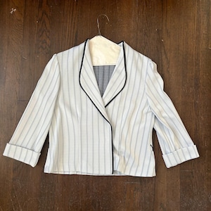 May include: A white blazer with vertical black pinstripes, a notched collar, and black trim. The blazer has three-quarter sleeves with folded cuffs. The garment is hanging on a hanger against a dark wood background.