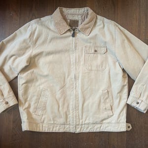 May include: A beige canvas jacket featuring a corduroy collar, a full-length front zipper, and long sleeves with buttoned cuffs. The garment includes a single chest pocket with a buttoned flap and two lower welt pockets, finished with a straight hem.