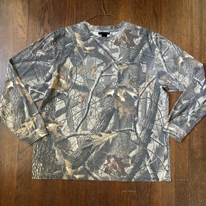 Puede incluir: Una camiseta de manga larga con cuello redondo que presenta un patrón de camuflaje forestal realista en tonos marrón, beige y gris, con imágenes de corteza de árbol, ramas y hojas de roble. Incluye un bolsillo en el pecho izquierdo.