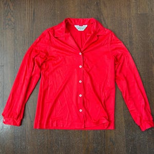 May include: A bright red button-down shirt with long sleeves. The shirt has a collar and white buttons down the front. The fabric appears to be a soft, flowing material. The shirt is displayed on a dark wood surface.
