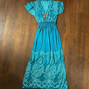 May include: A teal maxi dress with short sleeves and a V-neckline. The dress features a fitted waist and a flowing skirt. White paisley-like patterns adorn the sleeves and the bottom of the skirt. A decorative brooch is pinned at the neckline.