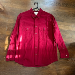 May include: A long-sleeved, button-down shirt in a deep red hue. The shirt features two chest pockets with button closures, a button-down collar, and buttoned cuffs. The Cabela's brand label is visible at the collar.