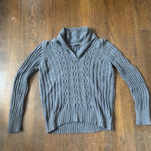 Puede incluir: Un suéter gris de punto trenzado con cuello chal. El suéter presenta un diseño acanalado en las mangas y el dobladillo inferior. La parte delantera tiene un patrón central de punto trenzado. La prenda está hecha de un material suave y cálido.