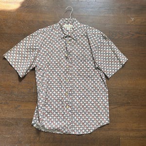 May include: Short-sleeved button-down shirt with a repeating pattern of red, white, and blue circles. The shirt is made of a lightweight fabric and has a collared neckline. The shirt is hanging on a metal hanger.