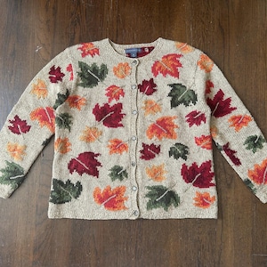 May include: Beige cardigan sweater with a pattern of colorful fall leaves in shades of red, orange, and green. The sweater has long sleeves and a button-up front. The leaves are scattered across the front and sleeves.