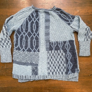 Könnte beinhalten: Ein Patchwork-Pullover in Blau- und Grautönen. Der Pullover weist verschiedene Strickmuster auf, darunter Zopfmuster und geometrische Designs. Die langen Ärmel haben gerippte Bündchen und der Ausschnitt ist rund.