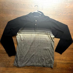 May include: Black long-sleeve polo shirt with a horizontal striped pattern in shades of gray and beige across the chest. The shirt has a collared neckline and is hanging on a white hanger.
