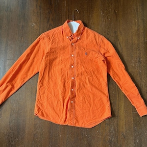 May include: An orange long-sleeved button-down shirt with a white collar. The shirt has a small blue logo on the chest. The shirt is hanging on a hanger.