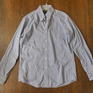 May include: A long-sleeved button-down shirt in a blue and white checkered pattern. The shirt is made of a lightweight fabric and features a button-down collar and cuffs. The shirt is hanging on a white hanger against a wooden background.