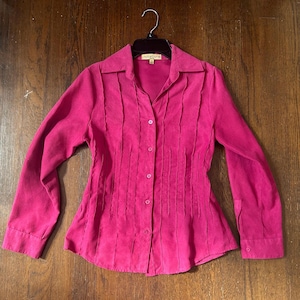 May include: A magenta-colored button-down shirt with long sleeves and a collared neckline. The shirt features vertical pleats down the front and is hanging on a black hanger. The fabric appears to be a soft, suede-like material.