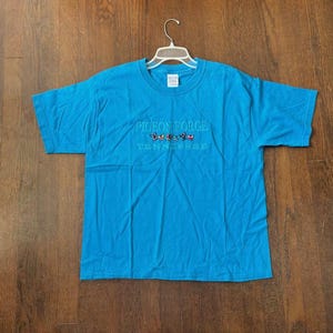 May include: A turquoise blue t-shirt with the embroidered text "PIGEON FORGE TENNESSEE" in white thread. The design includes colorful embroidered butterflies. The shirt is hanging on a white hanger against a brown wooden background.