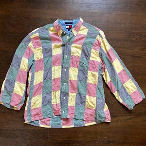 May include: A long-sleeved button-down shirt with a patchwork design. The shirt features a mix of red, yellow, green, and blue checkered patterns. The collar is blue, and the shirt has a small logo on the chest.