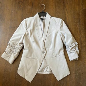 May include: A light beige blazer with three-quarter length, ruched sleeves. The blazer has a single-button closure and two front pockets. The jacket is hanging on a black hanger against a brown wooden surface.