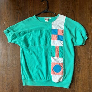 May include: A teal short-sleeved sweatshirt with a color block design. The color block features squares and a circle in shades of blue, coral, and white. The sweatshirt is hanging on a black hanger.