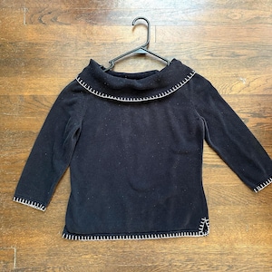 May include: A black sweater with a wide, folded collar and three-quarter sleeves. The collar, cuffs, and hem are trimmed with white stitching. The sweater is hanging on a black hanger.