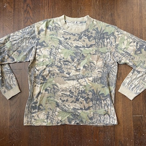 Könnte beinhalten: Ein langärmeliges T-Shirt mit einem Wald-Camouflage-Muster in Grün-, Braun- und Beigetönen. Das Shirt verfügt über einen Rundhalsausschnitt, lange Ärmel mit gerippten Bündchen und eine einzelne aufgesetzte Tasche auf der linken Brustseite.