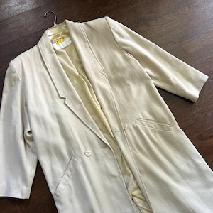 May include: A light beige blazer with a notched lapel, two-button closure, and front pockets. The longline jacket is hanging on a black hanger. The blazer has three-quarter sleeves and a light beige lining.