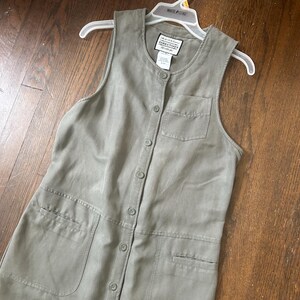 May include: A sleeveless olive green button-down dress featuring a round neckline, a single chest pocket, and two large front pockets. The interior label reads Directives. The dress is displayed on a white plastic hanger against a wooden surface.