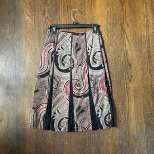 May include: A pleated skirt with a paisley pattern in shades of black, pink, and beige. The skirt has black vertical panels and is hanging on a black hanger against a wooden background. The skirt is a knee-length style.