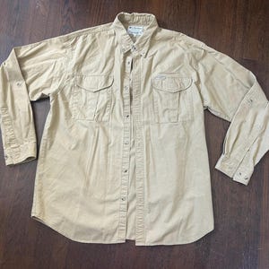 May include: A tan button-down shirt with two chest pockets and long sleeves. The shirt has a collared neckline and buttoned cuffs. The fabric appears to be a durable, woven material.