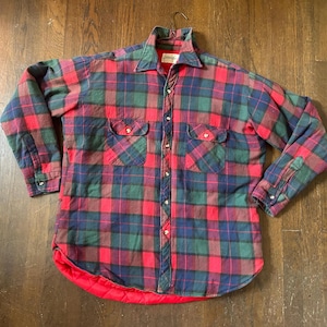 May include: A long-sleeve button-up shirt featuring a red, green, and navy blue plaid pattern. The garment includes a pointed collar, two chest pockets with button closures, buttoned cuffs, and a bright red quilted lining visible at the hem.