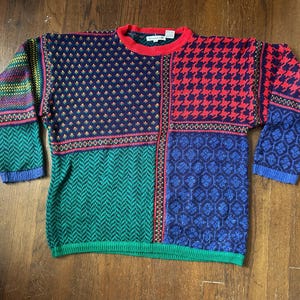 May include: A long-sleeve knit sweater featuring a patchwork design with various patterns including houndstooth, herringbone, and floral motifs in navy blue, red, green, and yellow. The sweater has a red crew neckline and ribbed cuffs.