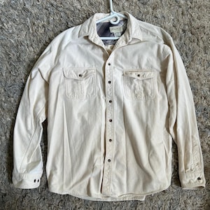 May include: A cream-colored long-sleeved button-down shirt with two chest pockets and a plaid flannel lining. The shirt has a button-up front and a relaxed fit.