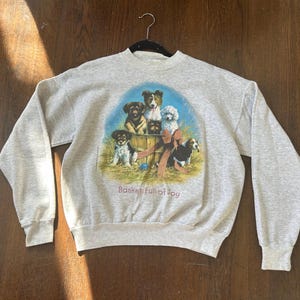 May include: A light gray sweatshirt featuring a colorful graphic of several dogs in a wooden basket. The phrase "Basket Full of Joy" is printed below the image. The sweatshirt has long sleeves and a crew neck.