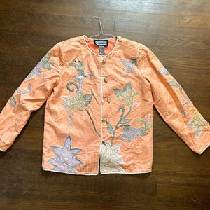 May include: Peach-colored jacket with floral embroidery. The jacket features long sleeves, a round neckline, and a button-up front. The floral design includes shades of green, lavender, and white. The jacket is hanging on a silver hanger.