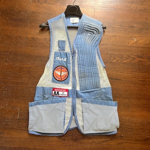 May include: Light blue shooting vest with mesh and pocket details. Features patches with text "Clark", "Winchester AA Straight", and a logo. The vest is hanging on a black hanger against a wooden background.