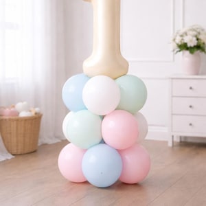 May include: A pastel-colored balloon arrangement for a first birthday celebration. A large, cream-colored number one balloon sits atop a pyramid of pastel pink, blue, green, and white balloons. The scene is set indoors with a light-colored wooden floor.