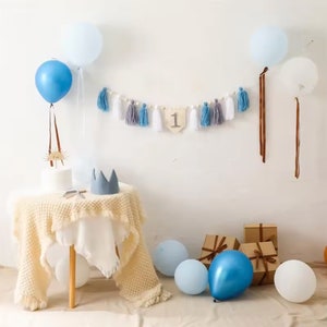 Puede incluir: Decoraci&oacute;n de fiesta de primer cumplea&ntilde;os con globos azules y blancos, una guirnalda de borlas y cajas de regalo. Una peque&ntilde;a mesa est&aacute; cubierta con una manta crema, y una corona azul est&aacute; encima. Tambi&eacute;n hay una tarta con un adorno en forma de sol.