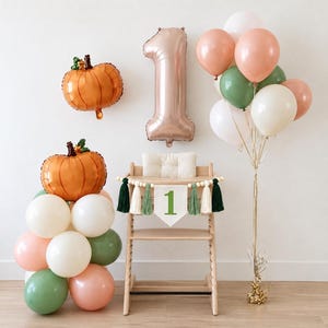 May include: A wooden high chair decorated with a banner featuring the number 1, surrounded by a balloon arrangement including orange pumpkin-shaped foil balloons, a large rose gold number 1 balloon, and a cluster of white, sage green, and peach balloons.