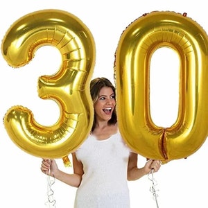 May include: Two large, gold foil balloons shaped as the numbers 3 and 0, forming the number 30. The balloons are held by a person wearing a white top. The balloons are inflated and have a shiny metallic finish, perfect for a birthday celebration.