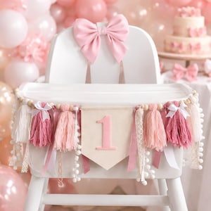 May include: A white high chair decorated for a first birthday celebration. It features a large pink bow, a banner with the number "1", tassels, and ribbons in shades of pink and white. The background includes balloons and a cake.