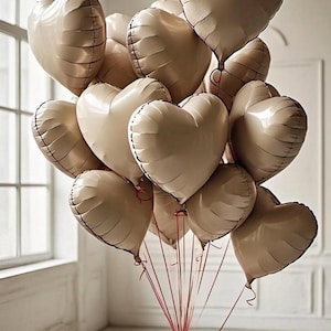 May include: A bouquet of beige heart-shaped balloons tied together with red ribbons. The balloons are floating in a room with a white wall and a window in the background.