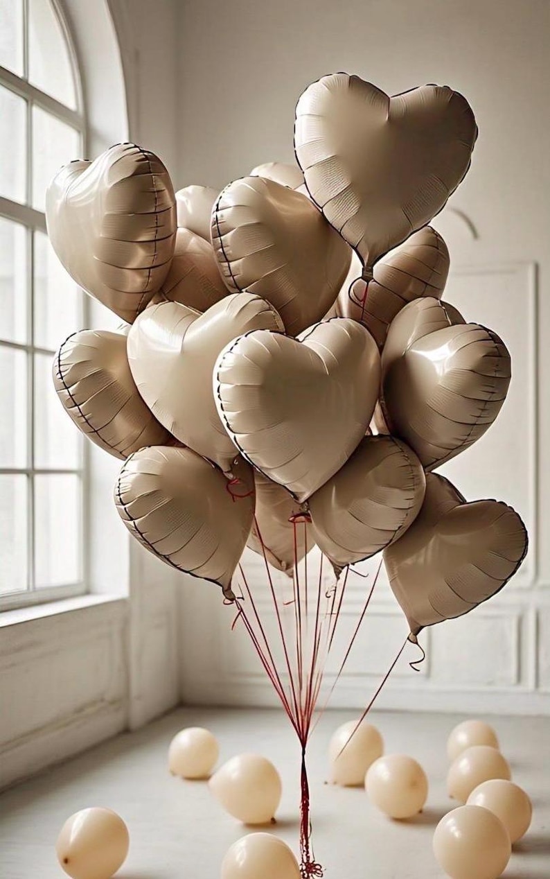 May include: A bouquet of beige heart-shaped balloons tied together with red ribbons. The balloons are floating in a room with a white wall and a window in the background.