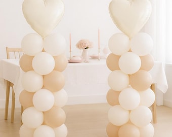 May include: Two vertical balloon columns standing on circular bases, each topped with a large cream-colored heart-shaped foil balloon. The columns are constructed from a mix of matte white and light beige latex balloons arranged in a stacked pattern.