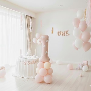 Op de afbeelding: Een kamer versierd voor een eerste verjaardag, met een grote ballon in de vorm van het cijfer één en groepen roze en witte ballonnen. Een kleine tafel is gedekt met een taart en een banner met de tekst "Happy Birthday".
