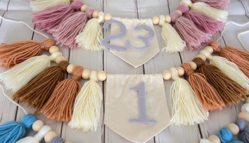 Puede incluir: Guirnaldas decorativas de borlas en varios colores, incluyendo rosa, crema, marr&oacute;n y azul. Cada guirnalda presenta cuentas de madera y banderines de tela con n&uacute;meros. Ideales para decoraciones de fiestas o decoraci&oacute;n del hogar.
