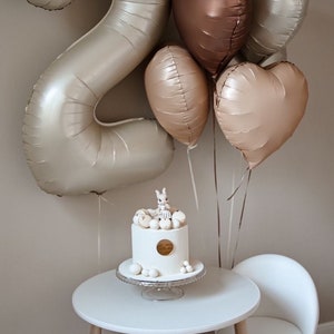 May include: A white cake with a bunny figurine on top, surrounded by white and gold balloons in the shape of hearts and the number two. The cake is on a white cake stand and a white table with a white chair.