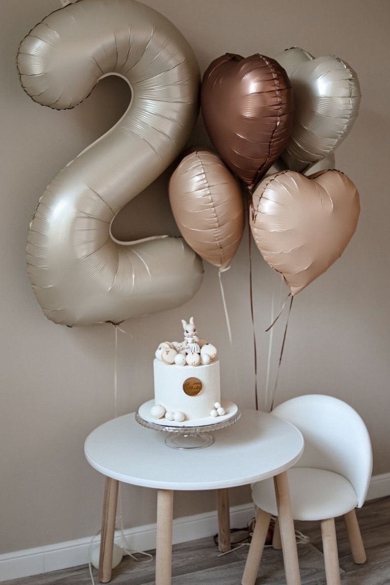 May include: A white cake with a bunny figurine on top, surrounded by white and gold balloons in the shape of hearts and the number two. The cake is on a white cake stand and a white table with a white chair.