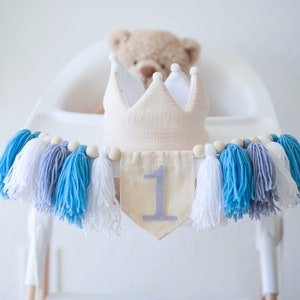 Puede incluir: Una corona color crema y una pancarta con el n&uacute;mero 1, adornada con borlas azules, blancas y grises. La pancarta es de tela beige y cuentas de madera. Un osito de peluche est&aacute; en el fondo.