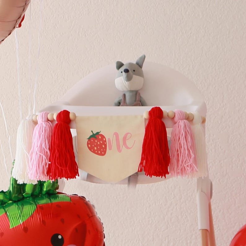 K&ouml;nnte beinhalten: Ein wei&szlig;er Hochstuhl mit einem Banner, auf dem "one" mit einer Erdbeergrafik steht. Das Banner ist mit rosa, roten und wei&szlig;en Quasten verziert. Ein graues Stofftier sitzt im Hochstuhl.